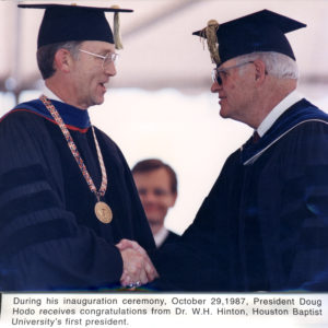 Two men in academic regalia shake hands and smile at an inauguration ceremony, making eye contact inside a university hall.