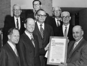 Group of men in suits, one holding a framed 60th Anniversary resolution, gather indoors during an HCU academic ceremony.