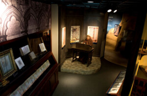 Exhibit for the 60th Anniversary displays a mannequin scribe at a wooden desk with illuminated manuscripts in an academic setting.