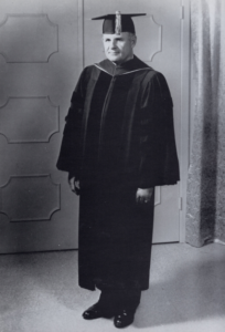 An older man in a cap and gown poses indoors for a formal 60th Anniversary graduation portrait, celebrating academic achievement.