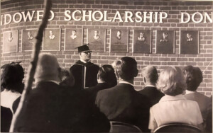 Individual in academic regalia addresses an audience beside the William H. Hinton donor wall at the Hinton Center.