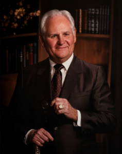 William H. Hinton smiling with glasses and cane, standing before bookshelves in an academic office setting.
