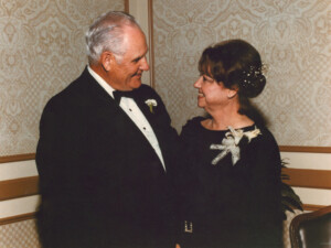 Elderly couple in formal attire exchange smiles in an ornate, warmly lit room during a ceremony honoring William H. Hinton.