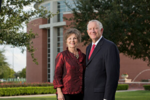 Smiling older couple in formal attire poses near a brick building and greenery on the HCU campus during a sunny day.