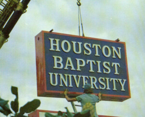 Worker stabilizes a large Houston Christian University sign as a crane lifts it on HCU campus under a cloudy sky.