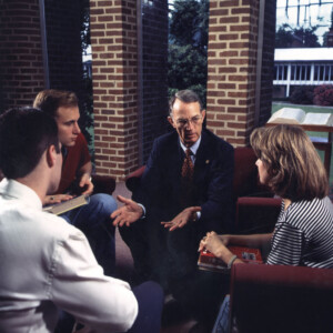 Students seated in a circle indoors, conversing with books nearby, likely in a group study or academic discussion setting.
