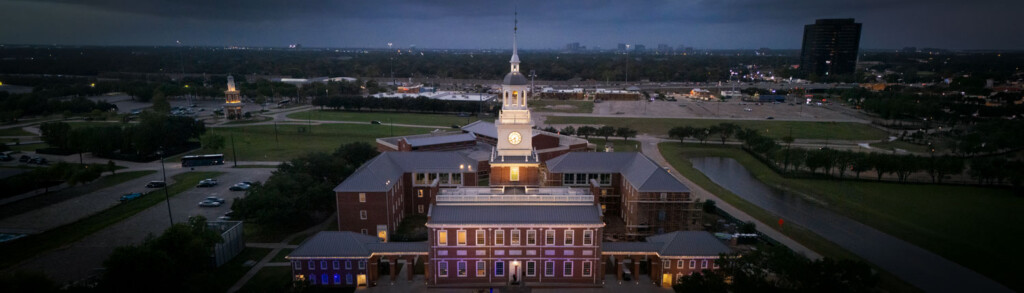 HCU campus aerial at dusk with Belin Tower clock glowing, city lights and surrounding trees visible.