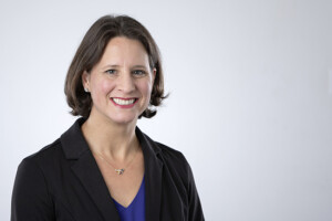 Confident woman with short brown hair in black blazer, Office of the Provost, light gray indoor background.