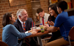 University President and three people share drinks, smiling and talking at a table inside an academic building.