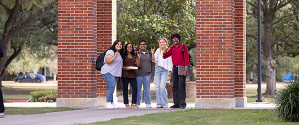 Five students smile together under a brick archway on HCU campus, with leafy trees in the background, enjoying student life.