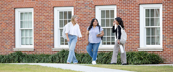 Three women walk and talk near a red brick building on the HCU campus, surrounded by greenery and enjoying student life together.