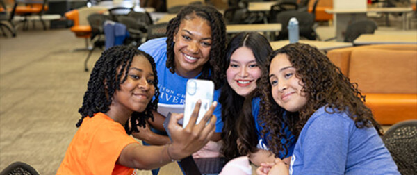 Four young women smile as they take a selfie in a lively campus lounge, surrounded by study tables and vibrant decor.
