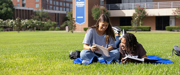 Students read and laugh together on the grass outside an HCU campus building, enjoying a sunny day.