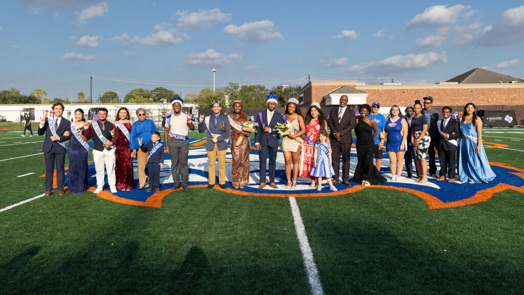 Formally dressed group poses in sunlight on HCU campus football field for Homecoming 2026, with stadium visible in background.