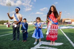 Students wearing sashes and crowns smile on HCU campus football field during a ceremony, posing for a celebratory photo.