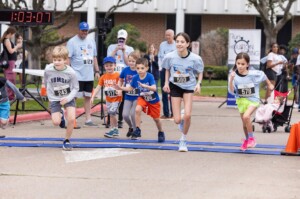 Six children start a race on HCU campus lawn, adults and a timer visible, campus buildings in the background.