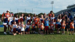 Cheerleaders, adults, and children smile with pom-poms on HCU campus football field, celebrating together.
