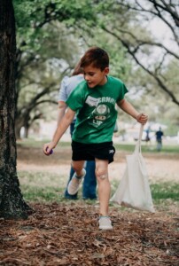Child in green shirt and shorts with a tote bag, walking near trees on HCU campus, bandage on knee visible.