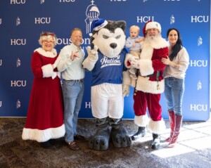 Santa, Mrs. Claus, four others, and a husky mascot smile together before an HCU backdrop at Houston Christian University event.
