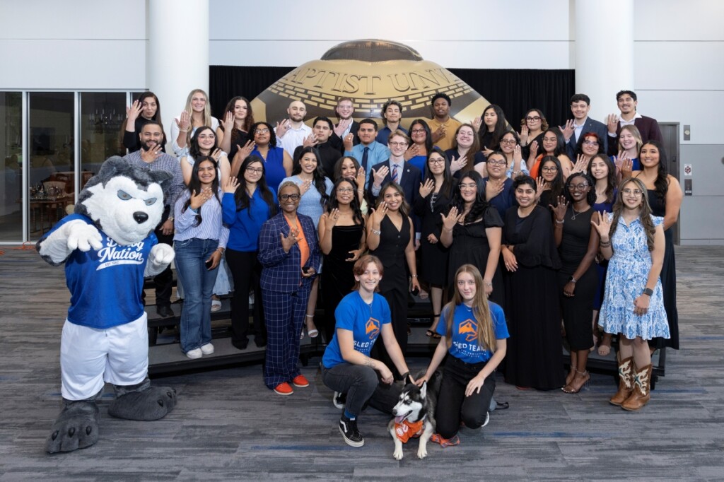 Students, staff, mascot, and a dog smiling together at an alumni event inside a university hall.