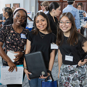 Three students with name tags and notebooks smile at a busy marketing event in an HCU campus building.