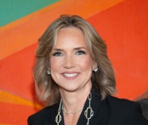 Woman with blond hair in a black blazer smiling at HCU Business Awards, colorful abstract backdrop behind her.