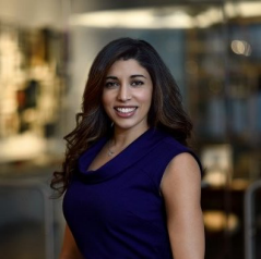 Nursing student with long dark hair in a sleeveless purple top smiles at an indoor HealthCare Lunch & Learn, blurred background.