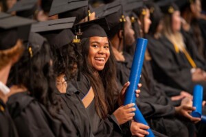 Houston Christian graduate in cap and gown smiles, holding blue diploma tube among classmates at commencement ceremony.