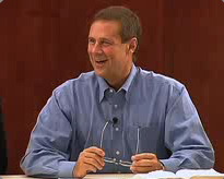 Sam Hunter in a blue shirt smiles at a table, holding glasses over an open book, engaged in academic study indoors.