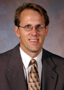 Mitchell J. Neubert in glasses, suit jacket, and patterned tie faces camera in academic setting with neutral indoor backdrop.