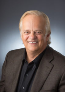 Dr. Bruce Hartman with light hair in a dark suit jacket and black shirt, posing against a neutral gray background indoors.