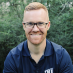 Man with glasses and red beard, smiling in a navy polo shirt outdoors on HCU campus with lush greenery in the background.