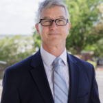 Smiling man in suit and glasses stands outdoors on HCU campus, with trees and sky behind him.