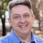 A smiling man in a blue collared shirt stands outside on the HCU campus with trees softly blurred in the background.