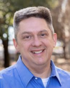 A smiling man in a blue collared shirt stands outside on the HCU campus with trees softly blurred in the background.