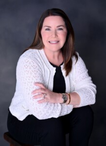 Female student in white sweater and black top smiles with arms crossed, seated indoors, likely during a campus academic activity.