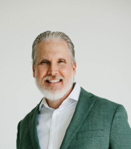Older man with gray hair and beard in green blazer, white shirt, and beige pants smiles against a neutral background.