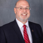 Professor with glasses, beard, and balding head in dark suit and red striped tie, smiling during lecture in academic setting.