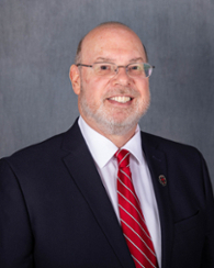 Professor with glasses, beard, and balding head in dark suit and red striped tie, smiling during lecture in academic setting.