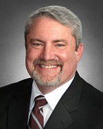 Gray-haired, bearded man in dark suit, member of Houston Christian University leadership, standing against a dark background.