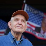 Jim McIngvale in brown cap and blue shirt stands before American flags at campus fundraiser; partially visible event banner in background.