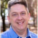 Smiling man with short gray hair in a blue shirt enjoys a business event outdoors on the HCU campus, trees softly blurred.