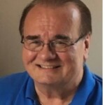 Smiling older man in glasses and blue shirt, wearing a gold necklace, prepares for mentoring conference indoors.