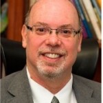 Business professor with glasses and beard in suit sits in academic office, prepared for panel discussion.