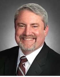 Professional man with gray hair and beard, dressed for business, smiling confidently against a dark backdrop.