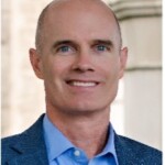Smiling bald man in blue blazer prepares to speak at mentoring event, stone wall softly blurred in the background.