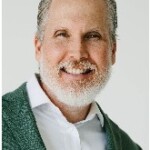 Older man with gray hair and beard, smiling, business attire, at academic panel discussion, facing the camera.