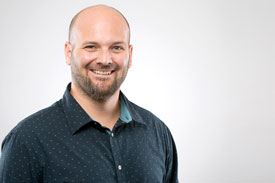 Jesse GrothOlson, smiling and bearded in a dark blue shirt, stands indoors before a light backdrop.