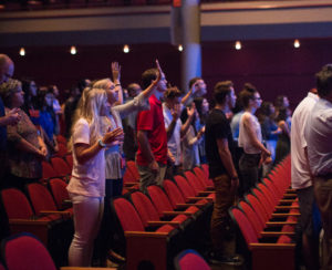 Worshipers with raised hands in a campus auditorium with red seats, illustrating Doing Good values during a church service.