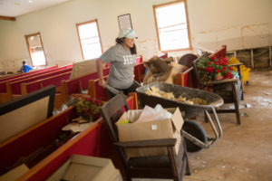 Woman cleaning a damaged church sanctuary; scattered pews, boxes, and wheelbarrow in an act of campus service and restoration.